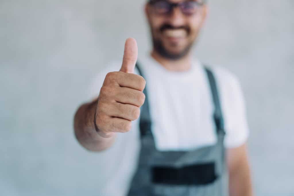 A shot of a confident, smiling professional HVAC technician in overalls and a white T-shirt, wearing a jumper outfit, standing in his workplace and showing a thumbs-up for One Hour Air Conditioning & Heating of Phoenix, AZ: Your HVAC System Partner.