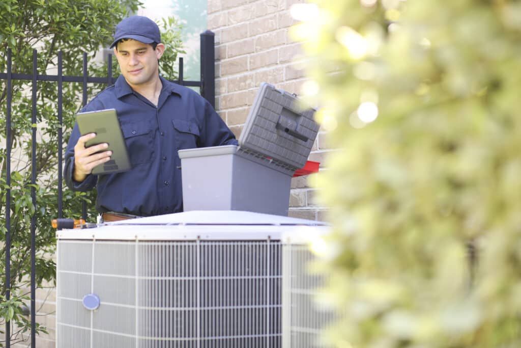 A man wearing a blue collar shirt is a residential HVAC systems repairman working at a residential home. He prepares to begin work by gathering appropriate tools and referring to a digital tablet.