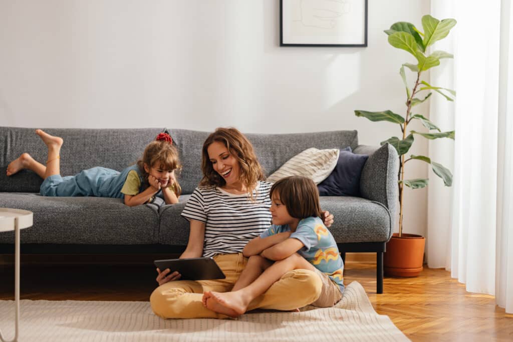 A beautiful woman is sitting on the floor with her kids, using a digital tablet to play games, watch movies, and relax at home, enjoying the cool and fresh air from the air conditioner. Her son is cuddled with her, while her daughter is lying on the sofa next to them. They are all smiling and looking down at the screen.